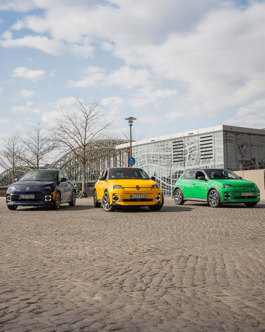 Drei Renault 5 E-Tech in Gelb, Dunkelblau und Grün in Kolonne auf einer Kölner Straße, im Vordergrund ein Frühlings-Blumenbeet
